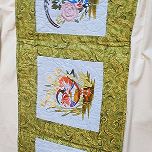 May include: A green fabric wall hanging with three white embroidered squares. Each square features a different embroidered bird design. The birds are surrounded by green foliage and flowers.