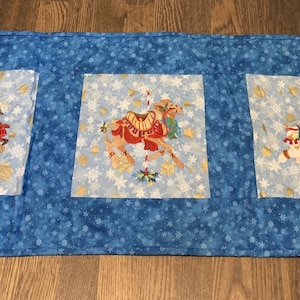 May include: A blue fabric table runner with three square panels featuring carousel horses. The horses are red, gold, and white, with colorful saddles and accents. The background is light blue with snowflakes.