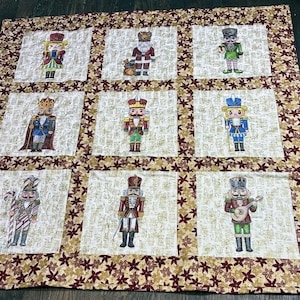 Könnte beinhalten: Ein quadratischer Quilt mit neun einzelnen Feldern, die jeweils eine einzigartige gestickte Nussknacker-Figur zeigen. Die Felder sind auf einem cremefarbenen Hintergrund mit goldener Textur angeordnet und von einer Bordüre aus dunkelroten und goldenen Blättern umrahmt.