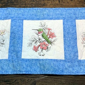 May include: A blue quilted table runner with three white squares featuring embroidered images of birds on branches with pink flowers. The birds are a bluebird, a hummingbird, and a robin.