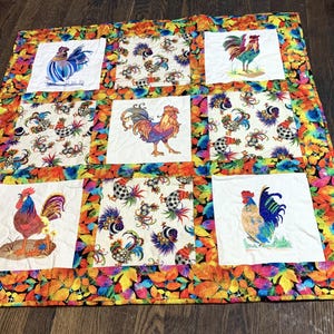 May include: A colorful quilt featuring embroidered rooster designs on white squares. The quilt has a vibrant border with autumn leaf patterns in shades of orange, yellow, and red. The roosters are depicted in various poses and colors.