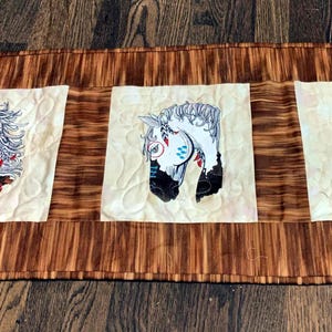 May include: A brown quilted table runner with three white squares featuring embroidered horse head designs. Each horse has a different colored bridle and mane.