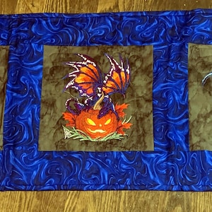 Może przedstawiać: Niebiesko-brązowy bieżnik na stół z haftowanymi wzorami smoków i dyni. Bieżnik ma niebieski wirujący wzór z trzema brązowymi kwadratami. Dwa kwadraty mają biały wzór smoka, a środkowy kwadrat ma smoka na dyni.