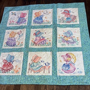 May include: A quilted blanket featuring nine square panels with embroidered illustrations of figures in bonnets and dresses. Each panel depicts a different activity, such as flying a kite, holding balloons, or drinking tea, set against a teal floral border.