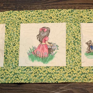 May include: A quilted wall hanging with three embroidered panels featuring girls in different scenes. The first panel shows a girl in a purple dress pushing a red wagon with a puppy. The second panel shows a girl in a pink dress holding a basket of flowers. The third panel shows a girl in a blue dress pouring tea for a teddy bear.