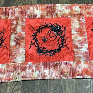 Puede incluir: Un camino de mesa rojo y blanco con tres cuadrados bordados en negro. Los cuadrados presentan una corona con una telaraña, una corona con cuervos y una corona con murciélagos.