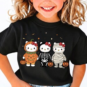 May include: A black short-sleeve t-shirt featuring a graphic of three white cat characters dressed in Halloween costumes: a pumpkin, a skeleton, and a mummy. Each character wears a red bow and holds a small orange pumpkin bucket for trick-or-treating.