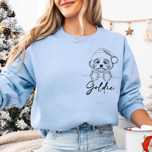 May include: Light blue sweatshirt featuring a black and white line drawing of a dog wearing a Santa hat. The dog's name, "Goldie," is written in cursive below the drawing. The sweatshirt is a solid light blue color.