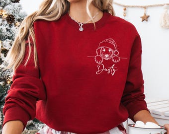 May include: A red sweatshirt featuring a white line drawing of a dog wearing a Santa hat. The dog's name, "Dusty," is written in cursive below the drawing. The sweatshirt is a solid red color.