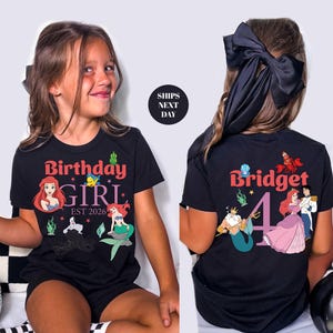 A young girl wearing a black t-shirt with a Disney character design and text, sitting against a white background.