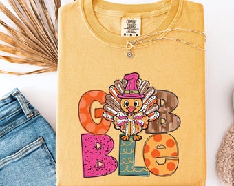 May include: A mustard yellow t-shirt featuring a graphic of a turkey wearing a pink hat and a polka dot bow tie. The word GOBBLE is printed in large, colorful, patterned letters surrounding the turkey illustration. The shirt has a Comfort Colors label.