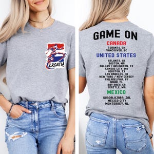 A woman wearing a grey t-shirt with a graphic of a person in a red and blue outfit, along with text that says "GAME ON CANADA" and a list of locations.