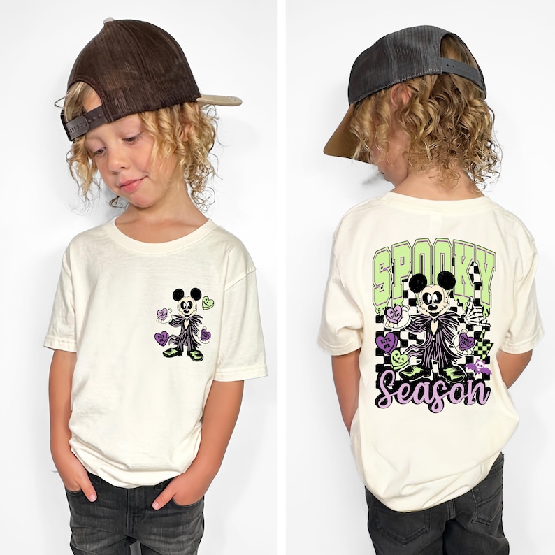 May include: Off-white short-sleeved t-shirt featuring a cartoon character with a Halloween theme. The front of the shirt has a small graphic, while the back displays the words "Spooky Season" with a larger design. The shirt is worn by a child.