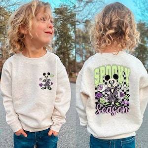 May include: A light beige sweatshirt featuring a Halloween-themed graphic. The front displays a cartoon character with hearts and flowers. The back of the sweatshirt has the words "Spooky Season" with a matching character and checkered pattern.