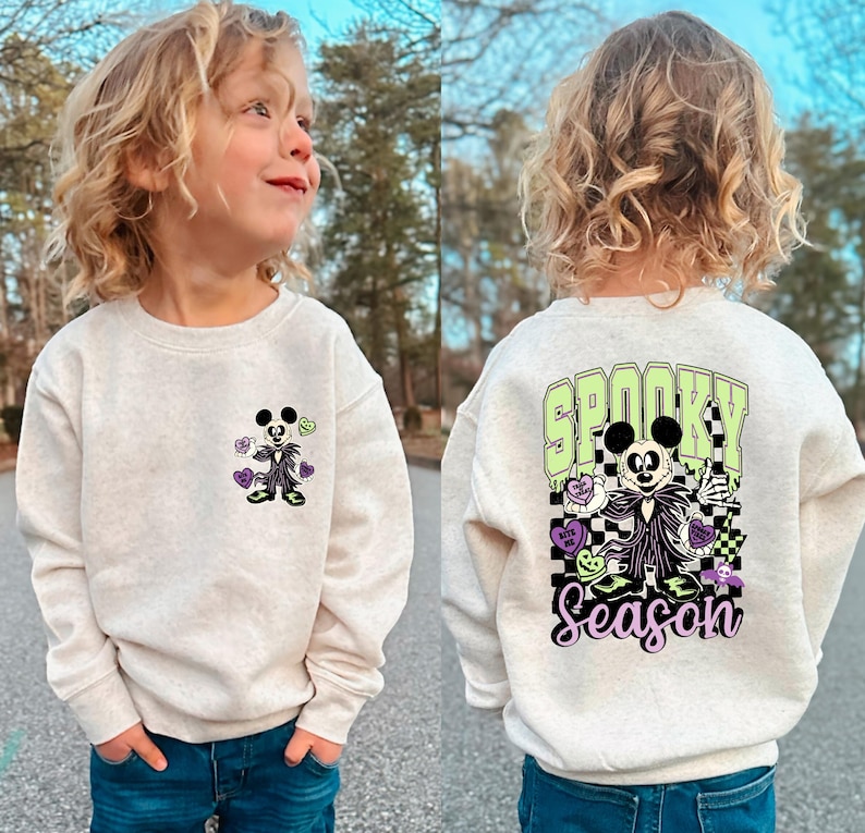 May include: A light beige sweatshirt featuring a Halloween-themed graphic. The front displays a cartoon character with hearts and flowers. The back of the sweatshirt has the words "Spooky Season" with a matching character and checkered pattern.