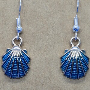 May include: A pair of silver seashell earrings with blue enamel detailing. The earrings have a hook closure and a small, round bead above the shell design. The shell design has a silver outline and a ribbed pattern.