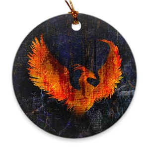 May include: A round ceramic ornament with a fiery orange phoenix rising from a dark blue and black background. The phoenix has a detailed design with orange and yellow feathers.