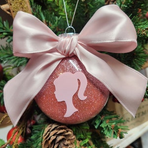 May include: A pink glitter Christmas ornament with a pink silhouette of a woman's head and a pink satin bow.