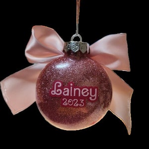 May include: Pink glitter Christmas ornament with a pink satin bow and the name "Lainey" and the year "2023" written on it.