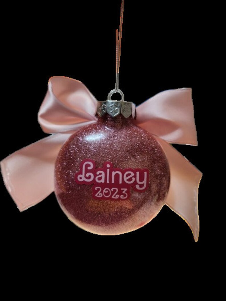 May include: Pink glitter Christmas ornament with a pink satin bow and the name "Lainey" and the year "2023" written on it.