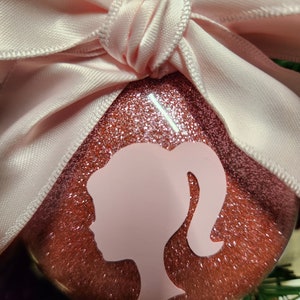 May include: A pink glitter Christmas ornament with a pink bow and a white silhouette of a woman's head and shoulders.