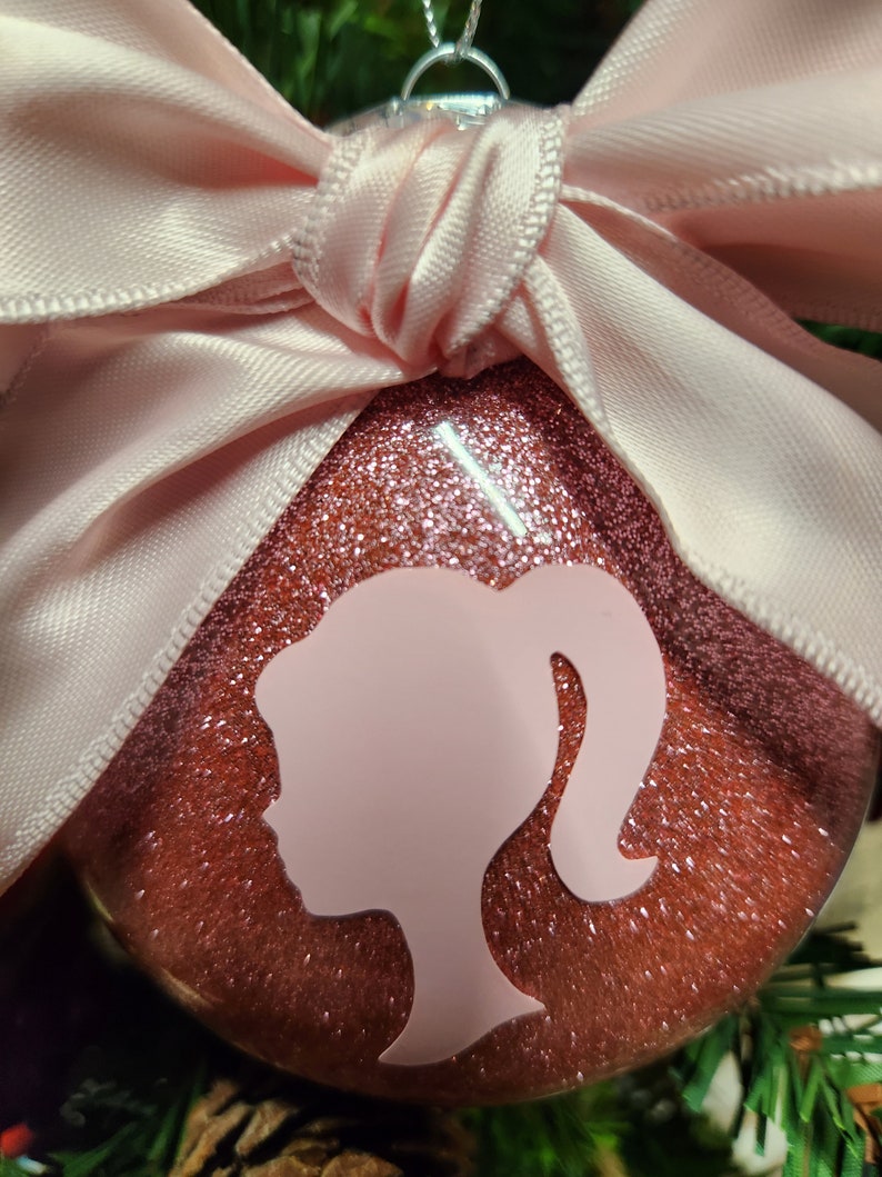 May include: A pink glitter Christmas ornament with a pink bow and a white silhouette of a woman's head and shoulders.