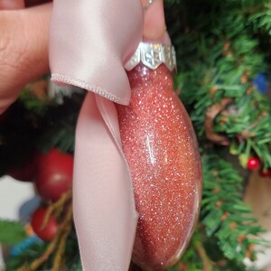 May include: A pink satin ribbon tied around a red glitter Christmas ornament. The ornament has a silver cap and is hanging from a silver string.