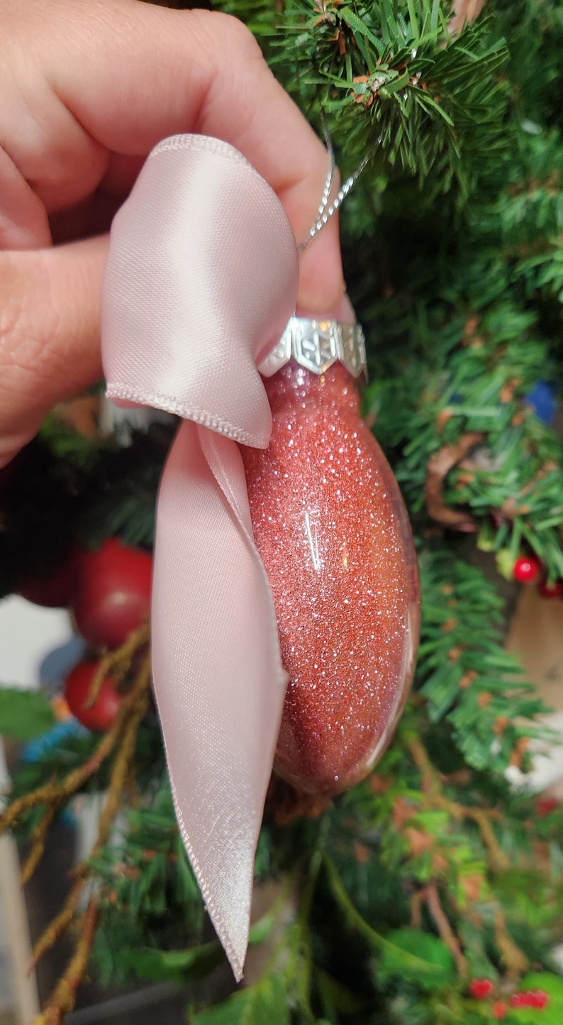 May include: A pink satin ribbon tied around a red glitter Christmas ornament. The ornament has a silver cap and is hanging from a silver string.