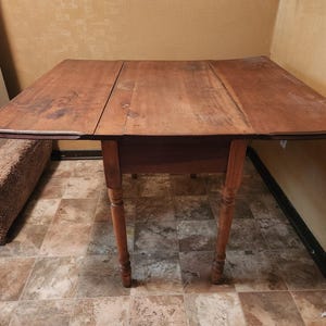 May include: A wooden drop-leaf table with a rectangular top and four turned legs. The table is made of a warm-toned wood, with visible grain and some wear. The table is approximately 36 inches wide when fully extended.