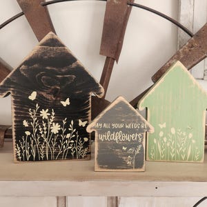 May include: Three wooden house-shaped decorations with floral and butterfly designs. The largest is black, the smallest is gray, and the third is green. The gray house has the text "MAY ALL YOUR WEEDS BE wildflowers".
