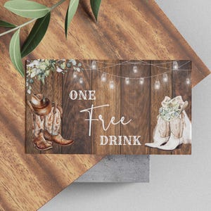 May include: A rustic-themed card with the words "ONE Free DRINK" on a wooden background. The card features illustrations of cowboy boots, a hat, and string lights. The card is decorated with greenery.