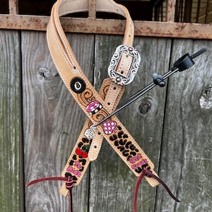 Puede incluir: Guarnición de caballo de cuero color canela con diseños decorativos pintados. La guarnición presenta una hebilla plateada, detalles en rojo y las frases "To Ride" y "Get Ready". El diseño incluye cerezas, dados y un patrón de cuadros rosas y blancos.