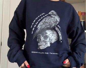 Puede incluir: Una sudadera azul marino de manga larga con un estampado gráfico blanco de un cuervo posado sobre un cráneo humano. El texto dice: "take thy beak from out my heart, and take thy form from off my door!" "NEVERMORE." y "EDGAR ALLAN POE - THE RAVEN."