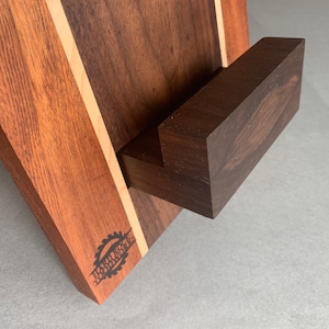 Puede incluir: Un soporte de tel&eacute;fono de madera con un acabado marr&oacute;n oscuro y una franja marr&oacute;n claro. El soporte tiene una peque&ntilde;a repisa rectangular para sostener un tel&eacute;fono. El soporte est&aacute; grabado con las palabras "Harley's Woodworks".
