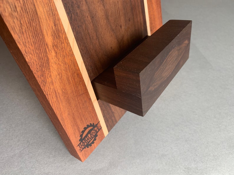 Puede incluir: Un soporte de tel&eacute;fono de madera con un acabado marr&oacute;n oscuro y una franja marr&oacute;n claro. El soporte tiene una peque&ntilde;a repisa rectangular para sostener un tel&eacute;fono. El soporte est&aacute; grabado con las palabras "Harley's Woodworks".