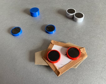 Puede incluir: Primer plano de un proyecto artesanal con dos círculos rojos y negros unidos a un trozo de tela. La tela es beige y tiene un borde de cinta marrón. También hay varios círculos azules y blancos esparcidos alrededor de la tela.