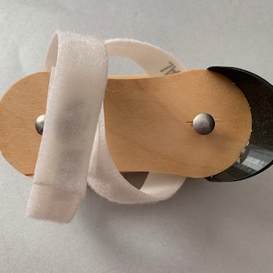 May include: A white fabric strap wraps around a wooden sandal with a black rubber toe. The sandal has two silver metal rivets.