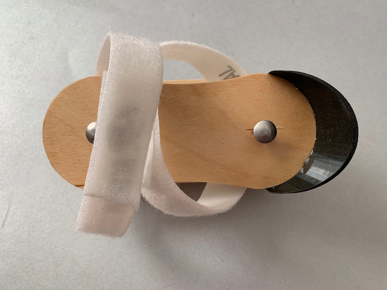 May include: A white fabric strap wraps around a wooden sandal with a black rubber toe. The sandal has two silver metal rivets.