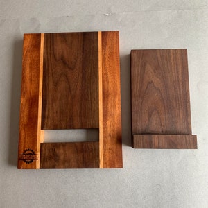 Puede incluir: Un soporte de tel&eacute;fono de madera con acabado de nogal. El soporte tiene una peque&ntilde;a ranura para cables de carga y una incrustaci&oacute;n decorativa de madera m&aacute;s clara. El soporte est&aacute; hecho por "Jardin Woodworks".