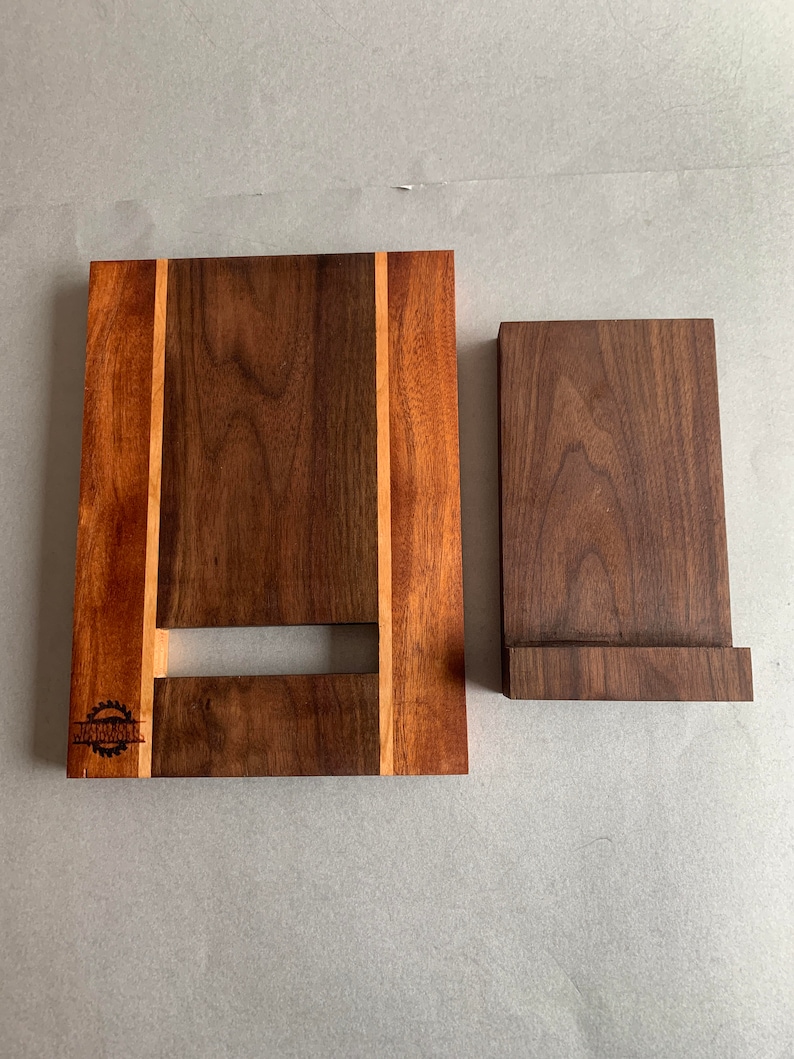Puede incluir: Un soporte de tel&eacute;fono de madera con acabado de nogal. El soporte tiene una peque&ntilde;a ranura para cables de carga y una incrustaci&oacute;n decorativa de madera m&aacute;s clara. El soporte est&aacute; hecho por "Jardin Woodworks".
