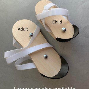 May include: Two pairs of wooden sandals with white straps. The larger pair is labeled "Adult" and the smaller pair is labeled "Child".  Larger size also available.