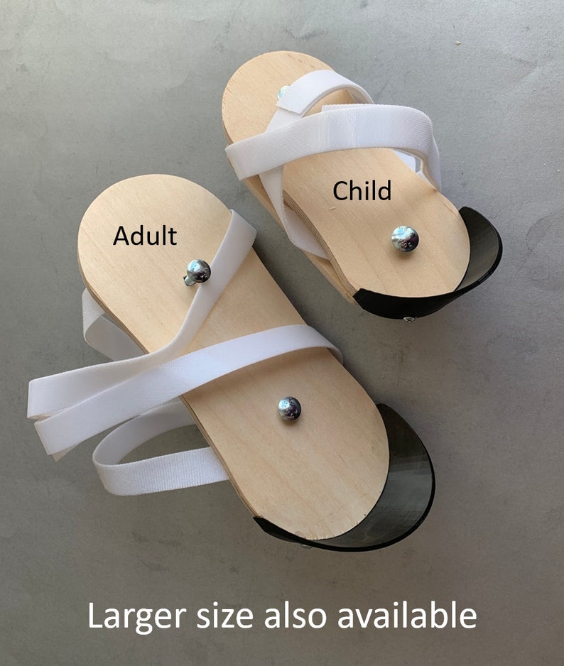 May include: Two pairs of wooden sandals with white straps. The larger pair is labeled "Adult" and the smaller pair is labeled "Child".  Larger size also available.