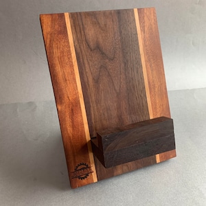 Puede incluir: Un soporte de tel&eacute;fono de madera con un dise&ntilde;o de madera de nogal y arce. El soporte tiene un peque&ntilde;o estante para sostener un tel&eacute;fono. El soporte est&aacute; grabado con las palabras "Woodworker's Den".