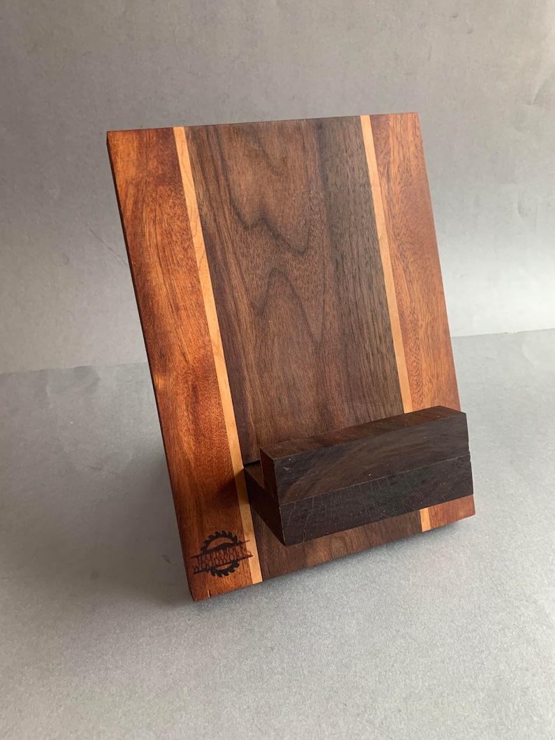 Puede incluir: Un soporte de tel&eacute;fono de madera con un dise&ntilde;o de madera de nogal y arce. El soporte tiene un peque&ntilde;o estante para sostener un tel&eacute;fono. El soporte est&aacute; grabado con las palabras "Woodworker's Den".