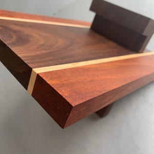Puede incluir: Una tabla de cortar de madera con una franja de color marr&oacute;n claro en el centro. La tabla est&aacute; hecha de madera de nogal y cerezo y tiene un acabado natural.
