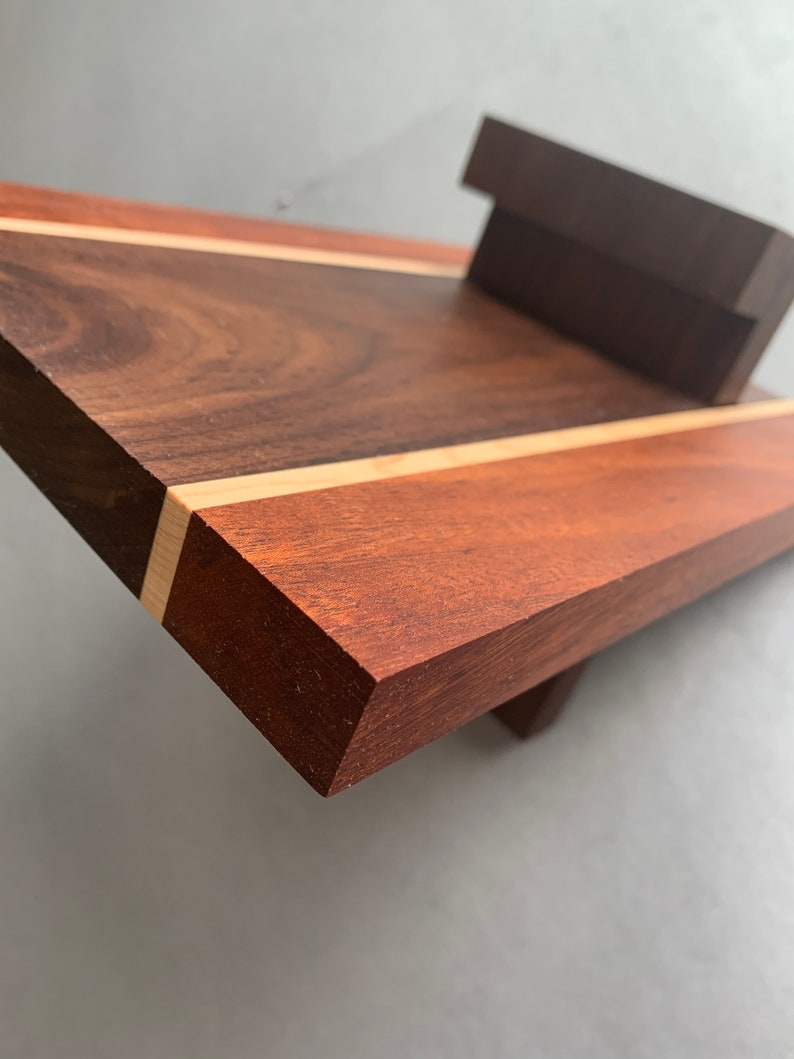 Puede incluir: Una tabla de cortar de madera con una franja de color marr&oacute;n claro en el centro. La tabla est&aacute; hecha de madera de nogal y cerezo y tiene un acabado natural.