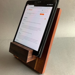 Puede incluir: Un soporte de tableta de madera con un acabado marr&oacute;n oscuro. El soporte est&aacute; hecho de dos piezas de madera, una de las cuales es un bloque rectangular y la otra es una pieza m&aacute;s peque&ntilde;a e inclinada. El soporte sostiene una tableta, que muestra una receta del sitio web Allrecipes.com.