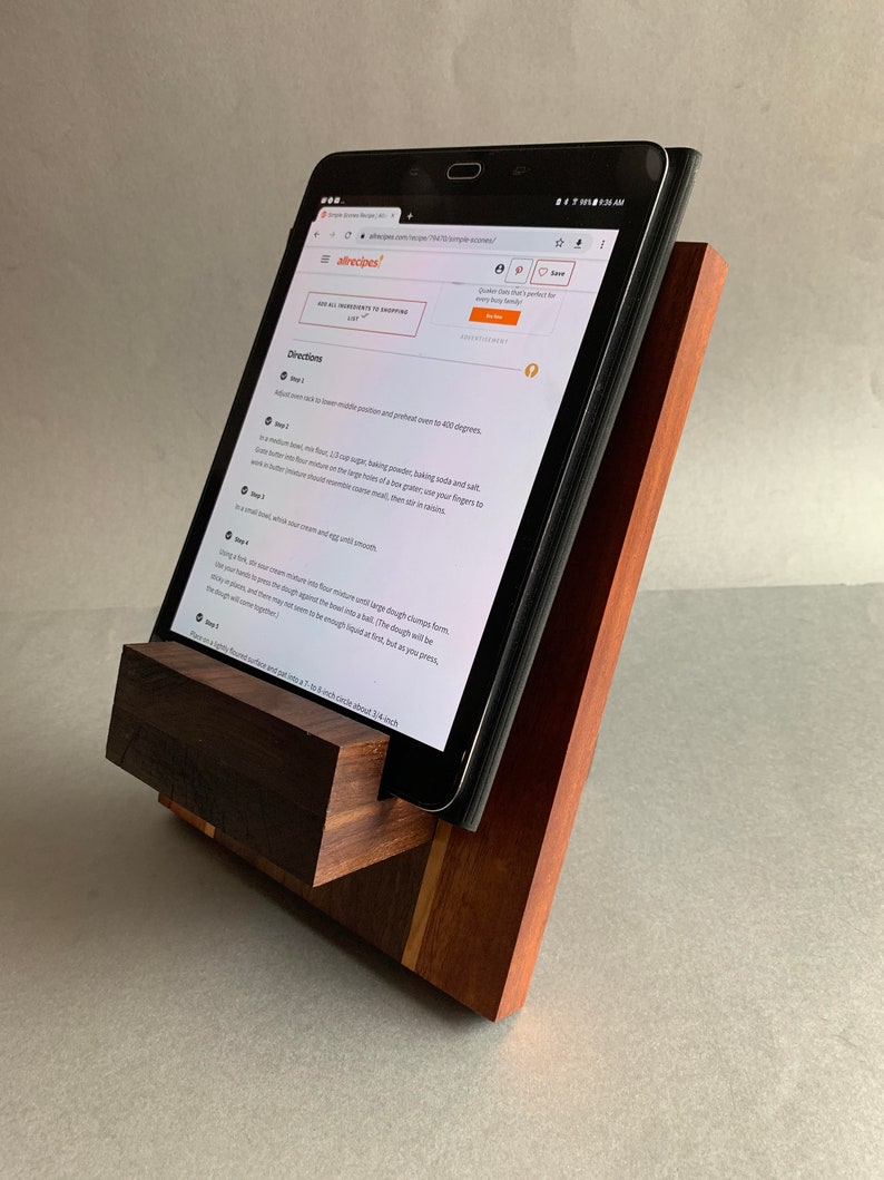 Puede incluir: Un soporte de tableta de madera con un acabado marr&oacute;n oscuro. El soporte est&aacute; hecho de dos piezas de madera, una de las cuales es un bloque rectangular y la otra es una pieza m&aacute;s peque&ntilde;a e inclinada. El soporte sostiene una tableta, que muestra una receta del sitio web Allrecipes.com.