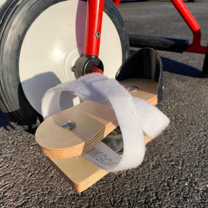 May include: A red tricycle with a white front wheel and a black rear wheel. The tricycle has a wooden footrest with a white strap attached to it.