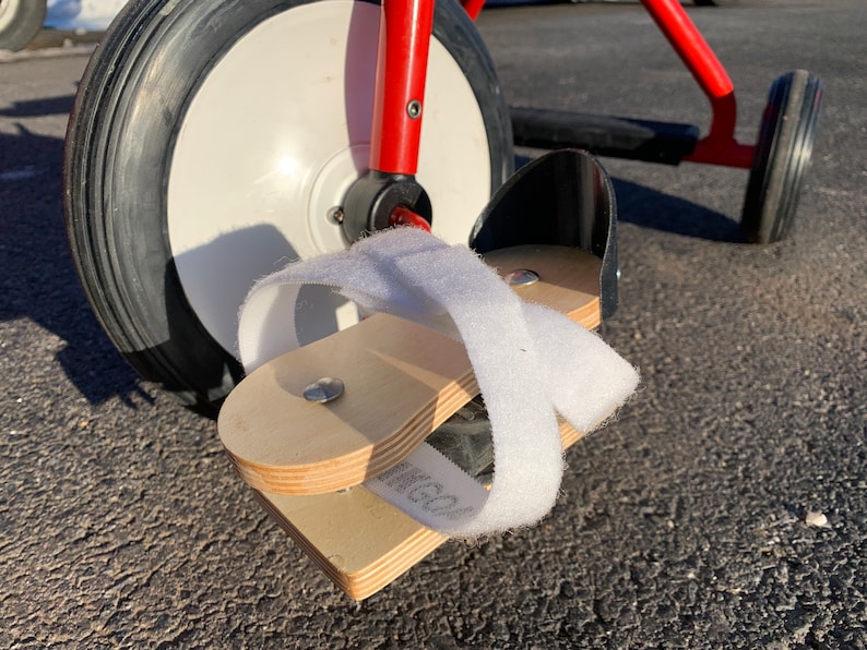 May include: A red tricycle with a white front wheel and a black rear wheel. The tricycle has a wooden footrest with a white strap attached to it.